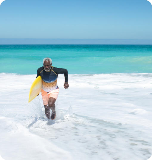 A senior man running through the surf on a tropic beach. He has a surfboard under his arm.