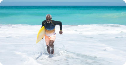 A senior man running through the surf on a tropic beach. He has a surfboard under his arm.