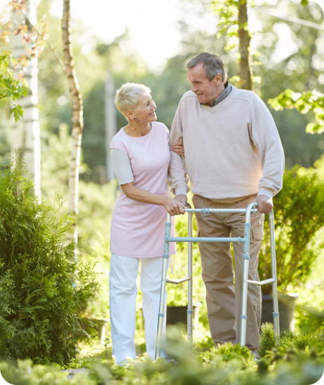 A senior man and woman outside on a sunny day. The man is leaning on a walker, and the woman is holding his arm affectionately. There are a lot of trees and other greenery in the background.