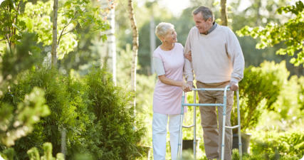 A senior man and woman outside on a sunny day. The man is leaning on a walker, and the woman is holding his arm affectionately. There are a lot of trees and other greenery in the background.