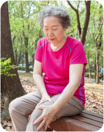 Photo of a senior woman sitting on a bench next to a walking trail in a park on a sunny day. She is holding her knee with a pained expression on her face.