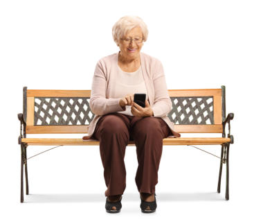 Photo of a senior woman on a white background sitting on a bench. She is smiling while she looks at her phone.