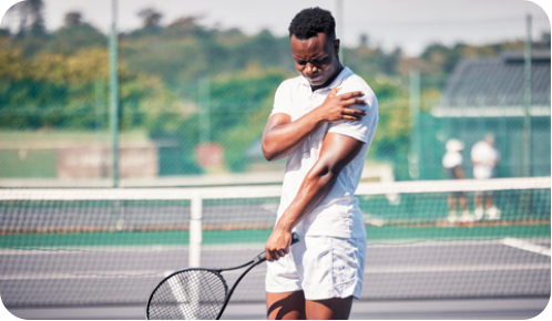 A young man is standing on a tennis court holding his shoulder and grimacing in pain. In his other hand he is holding a tennis racquet.